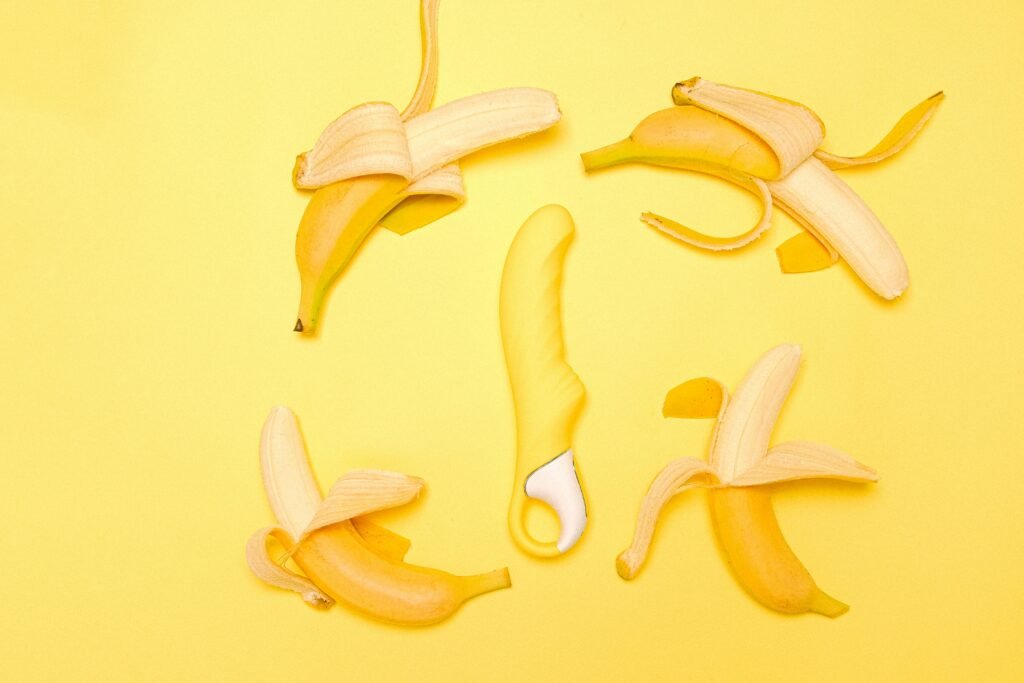 Creative flat lay of a yellow silicone vibrator surrounded by peeled bananas on a yellow background.